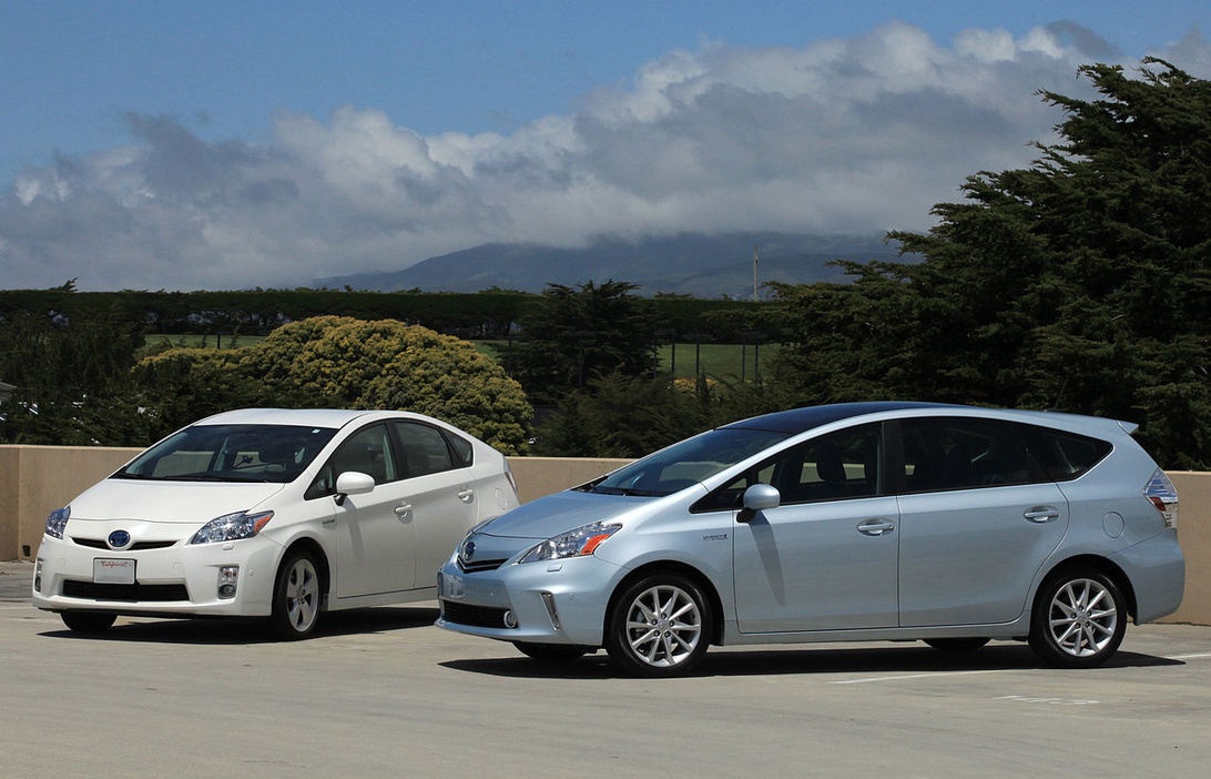 Två moderna Toyota Prius hybridbilar, en vit och en silverfärgad, parkerade bredvid varandra framför ett bergslandskap, vilket illustrerar miljövänliga transportalternativ för familjer.